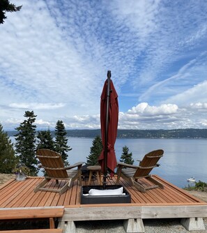 Un pontile in legno con due poltrone Adirondack e un ombrello rosso, affacciato su un lago tranquillo con una riva alberata e montagne in lontananza.