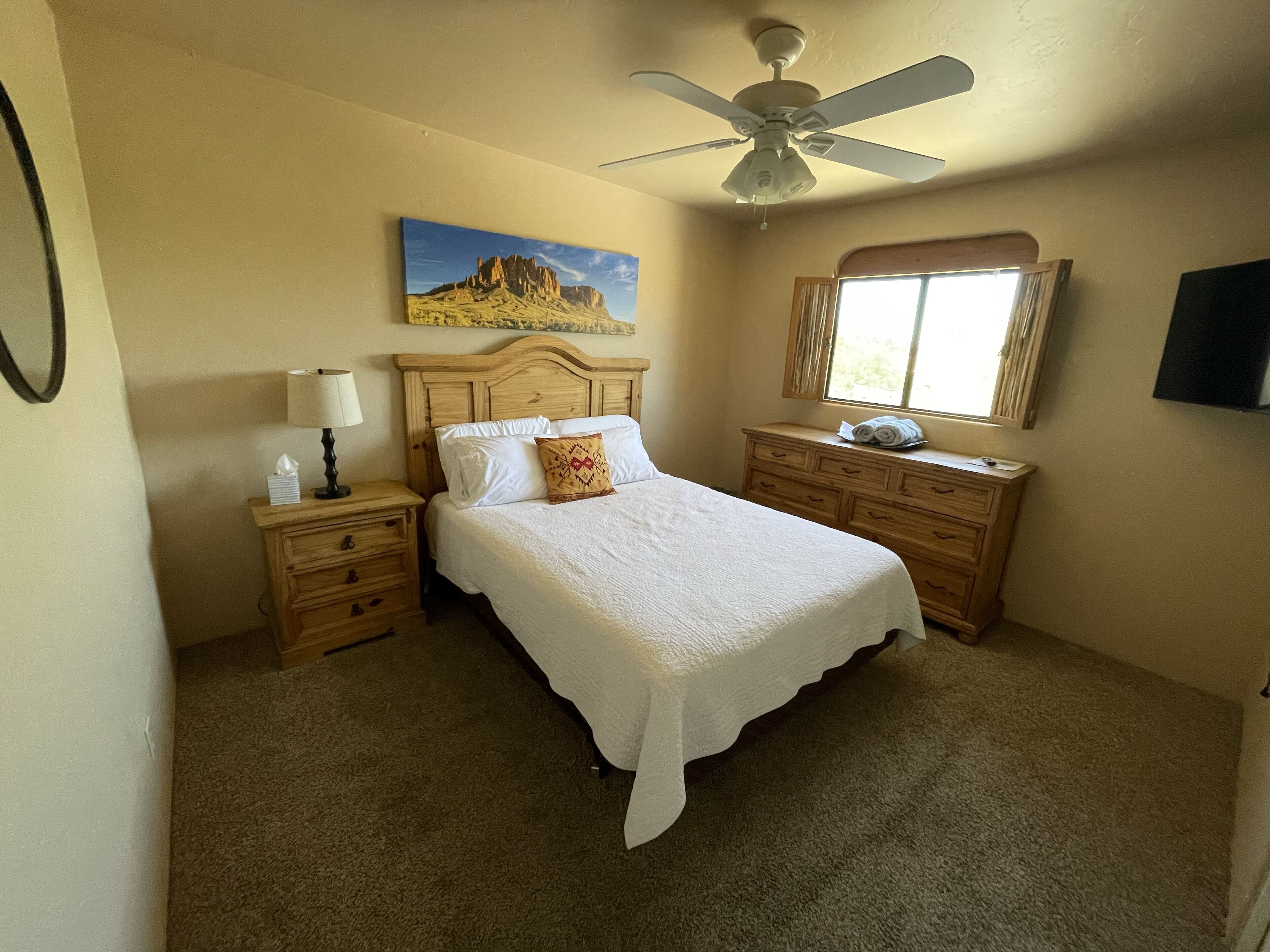 A bedroom with a bed, wooden nightstands, a ceiling fan, a window with curtains, and a painting on the wall.