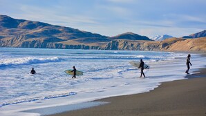 Une plage avec des vagues, des gens tenant des planches de surf et des montagnes qui se dessinent à l’horizon.