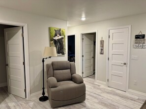 Une pièce avec un fauteuil beige, un lampadaire et un mur orné d’un tableau représentant un ours. On y trouve des portes blanches et un panneau sur le mur.