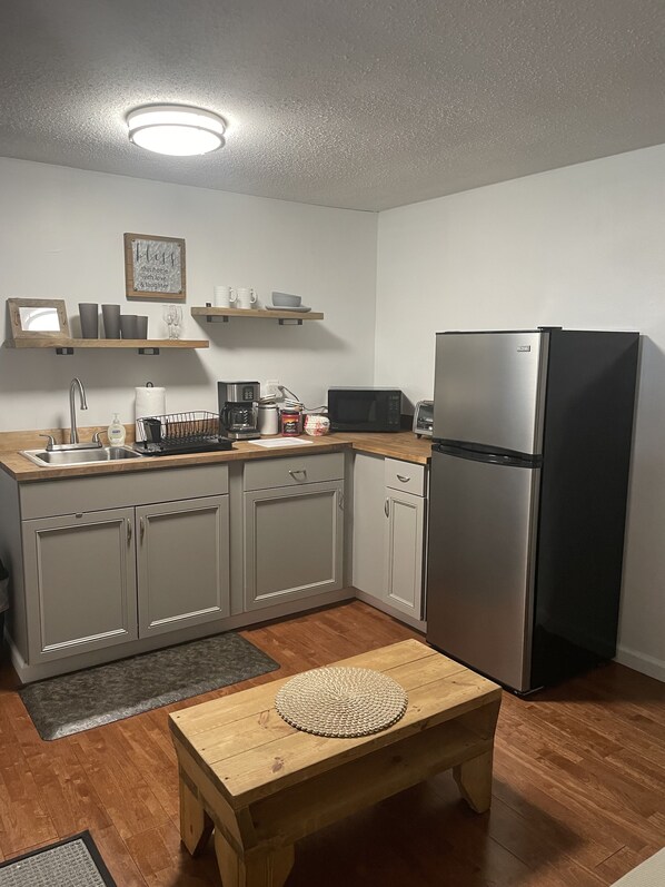 A compact kitchen with a sink, microwave, toaster, and refrigerator. There are wooden shelves above the counter and a small table with a woven placemat.