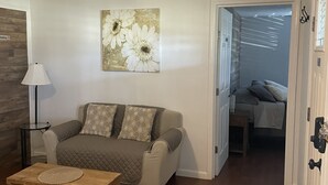 Een woonkamer met een bank, een salontafel en een schilderij aan de muur. Een deur leidt naar een andere kamer.