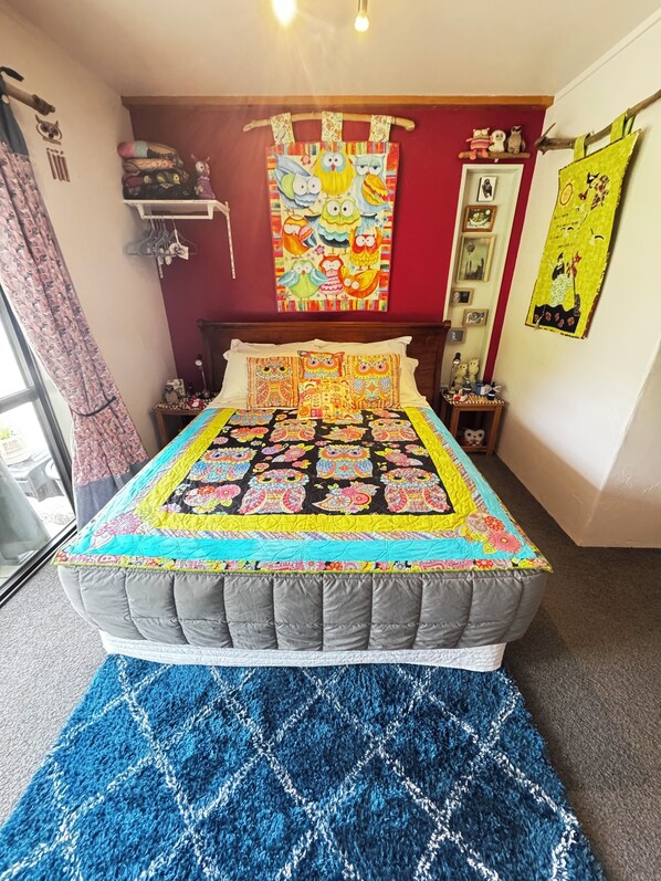 A bedroom with a bed covered in a colorful quilt, a red wall with a hanging tapestry, and a blue rug.
