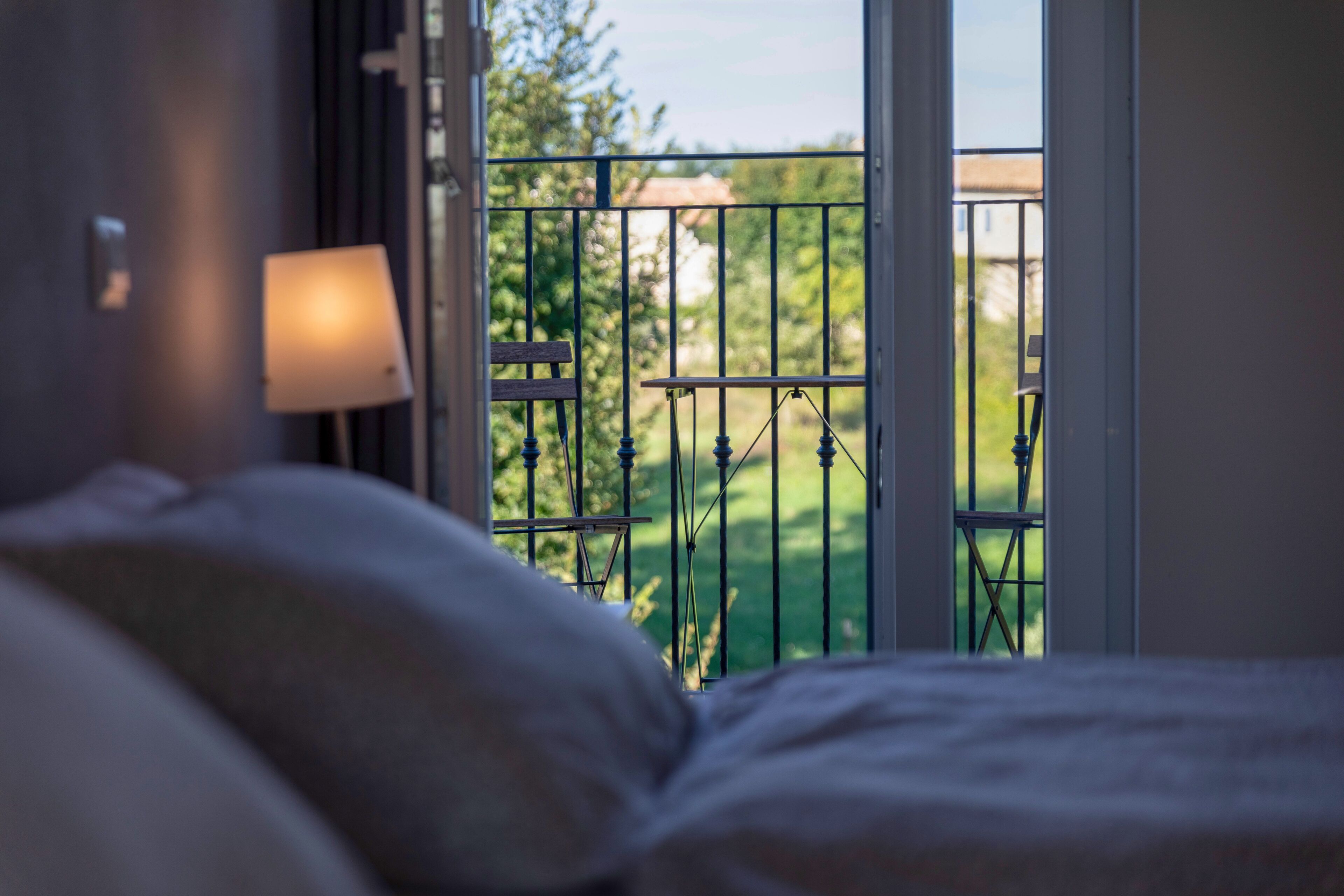 A bedroom with a bed, a nightstand, and a window with a view of a balcony and greenery.