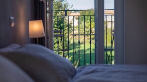 A bedroom with a bed, a nightstand, and a window with a view of a balcony and greenery.