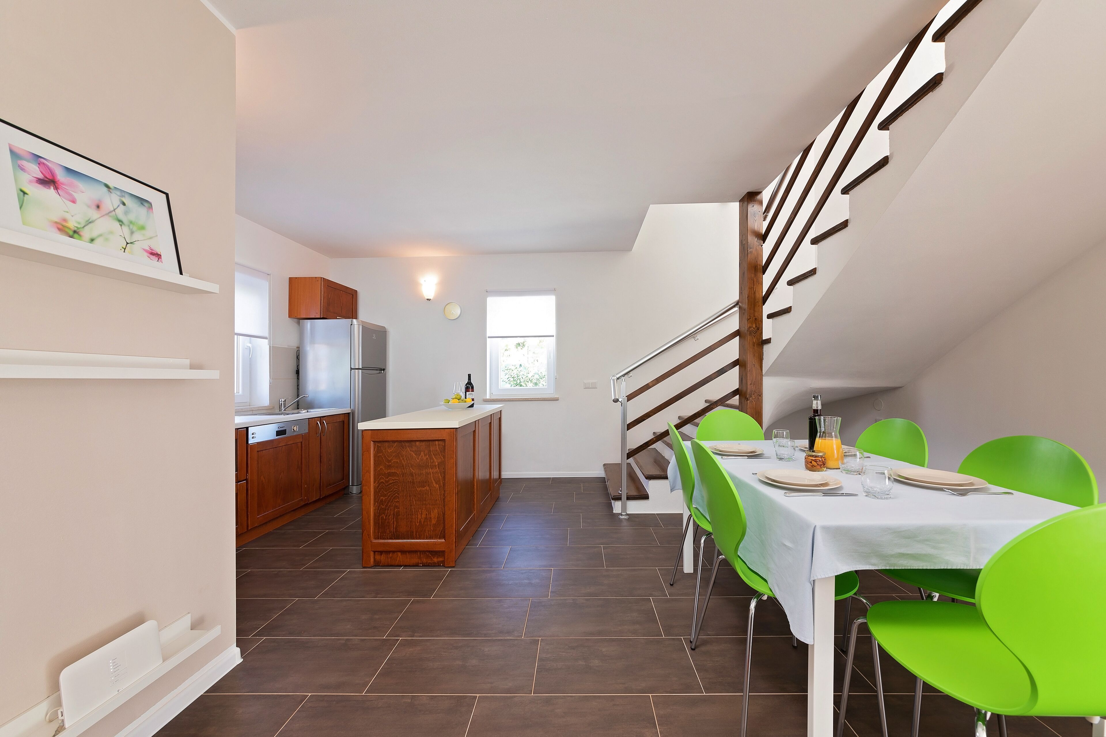 A modern kitchen with wooden cabinets, a built-in island, and a dining area with green chairs and a white table.