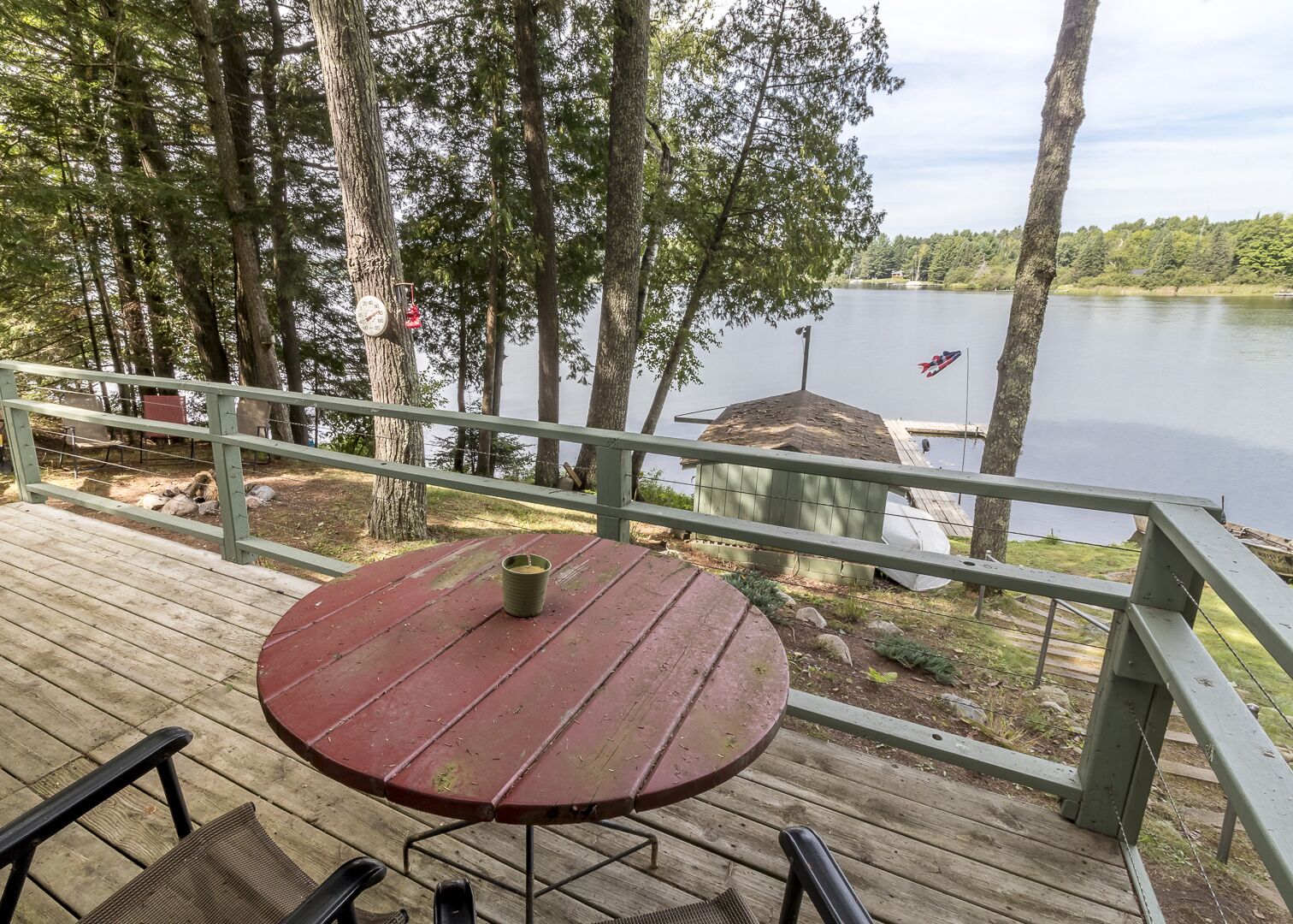 Une terrasse en bois avec une table ronde et des chaises, donnant sur un lac bordé d’arbres et sur un quai.