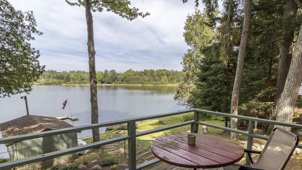 Une terrasse en bois avec une table et des chaises, donnant sur un lac et entourée d’arbres.