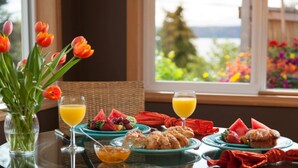 Une table de petit-déjeuner avec des fruits, des viennoiseries et du jus d’orange, placée devant une fenêtre donnant sur un jardin.