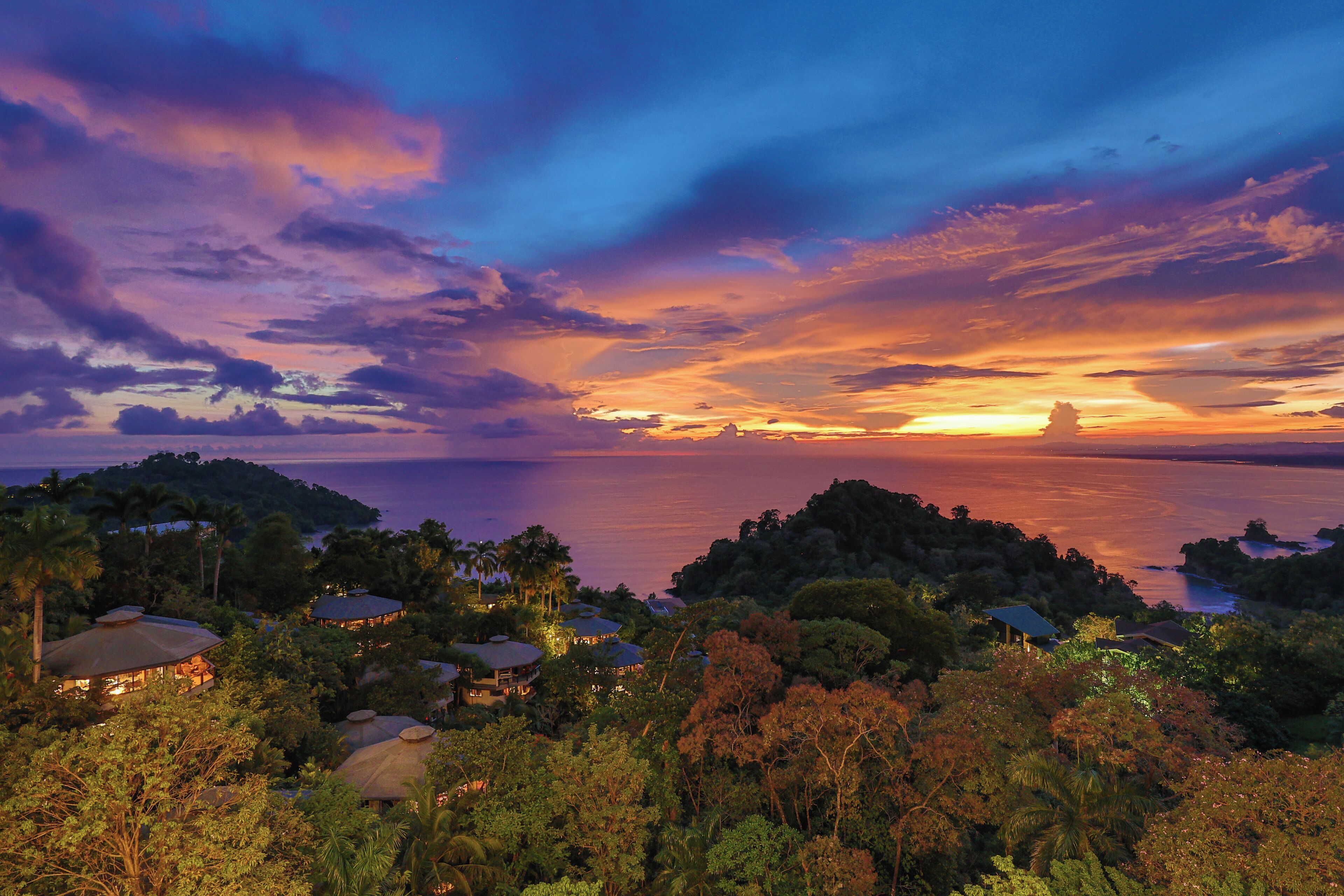 A coastal landscape with a sunset, houses surrounded by dense vegetation, and a calm sea.