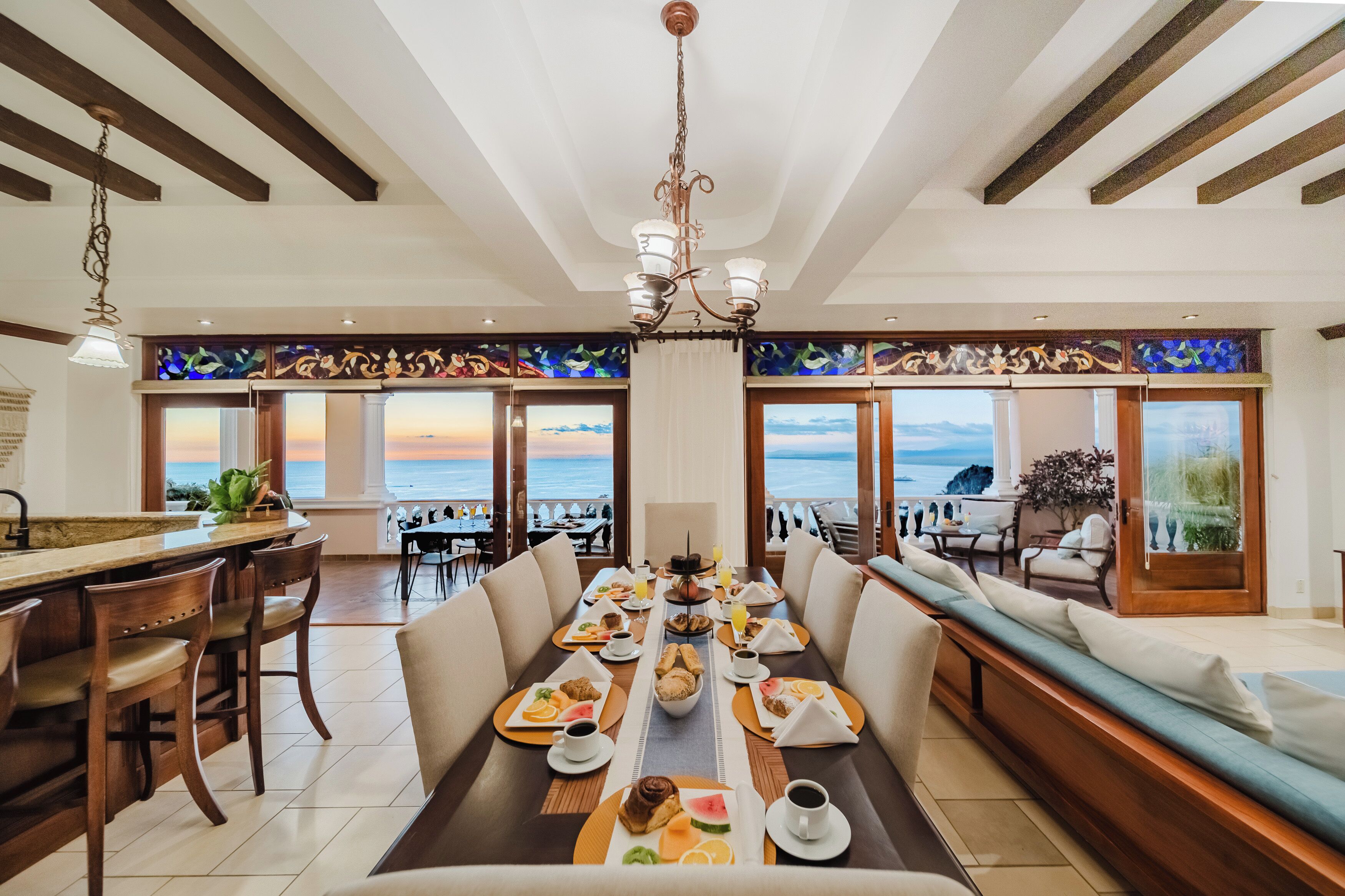 A dining area with a long table set for breakfast, large windows with ocean views, and a chandelier.