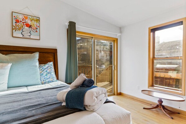 A neatly made bed with pillows and a rolled towel, a wooden headboard, a small round table, and a large window with a view of a wooden deck.