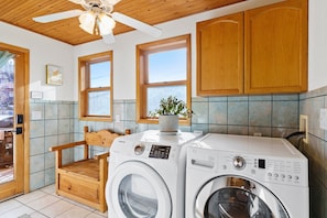A laundry room with two washing machines, wooden cabinets, a ceiling fan, and a window with a plant.