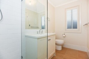 Un bagno con box doccia in vetro, lavandino bianco, wc e finestra con persiane.