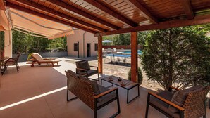 A covered outdoor seating area with lounge chairs and a table, a pool in the background, and a view of trees and a building.