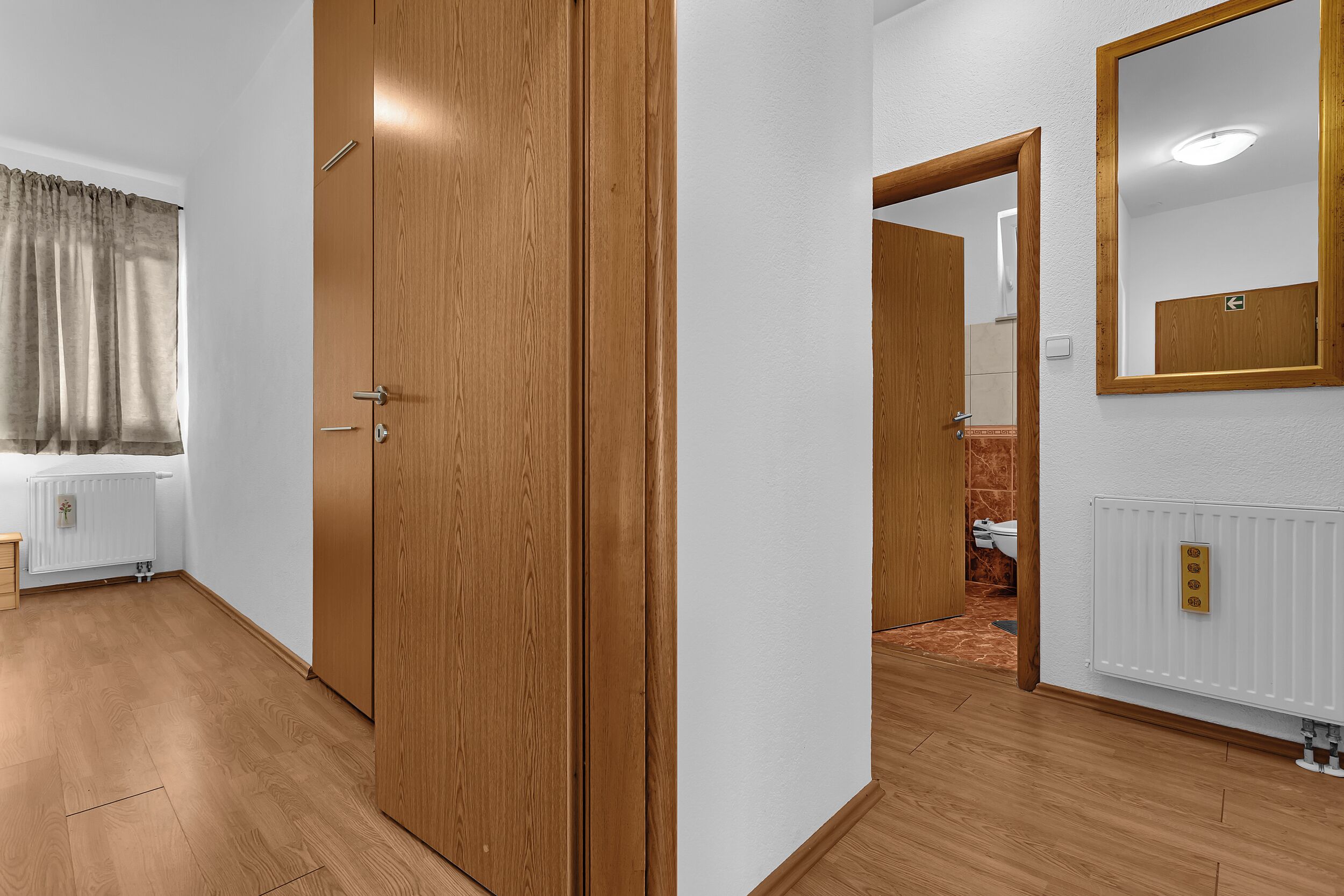 Une pièce avec un plancher en bois, un radiateur blanc, une armoire en bois et un miroir au mur.