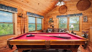 Un intérieur en bois avec une table de billard, un ventilateur de plafond et une fenêtre équipée de stores.
