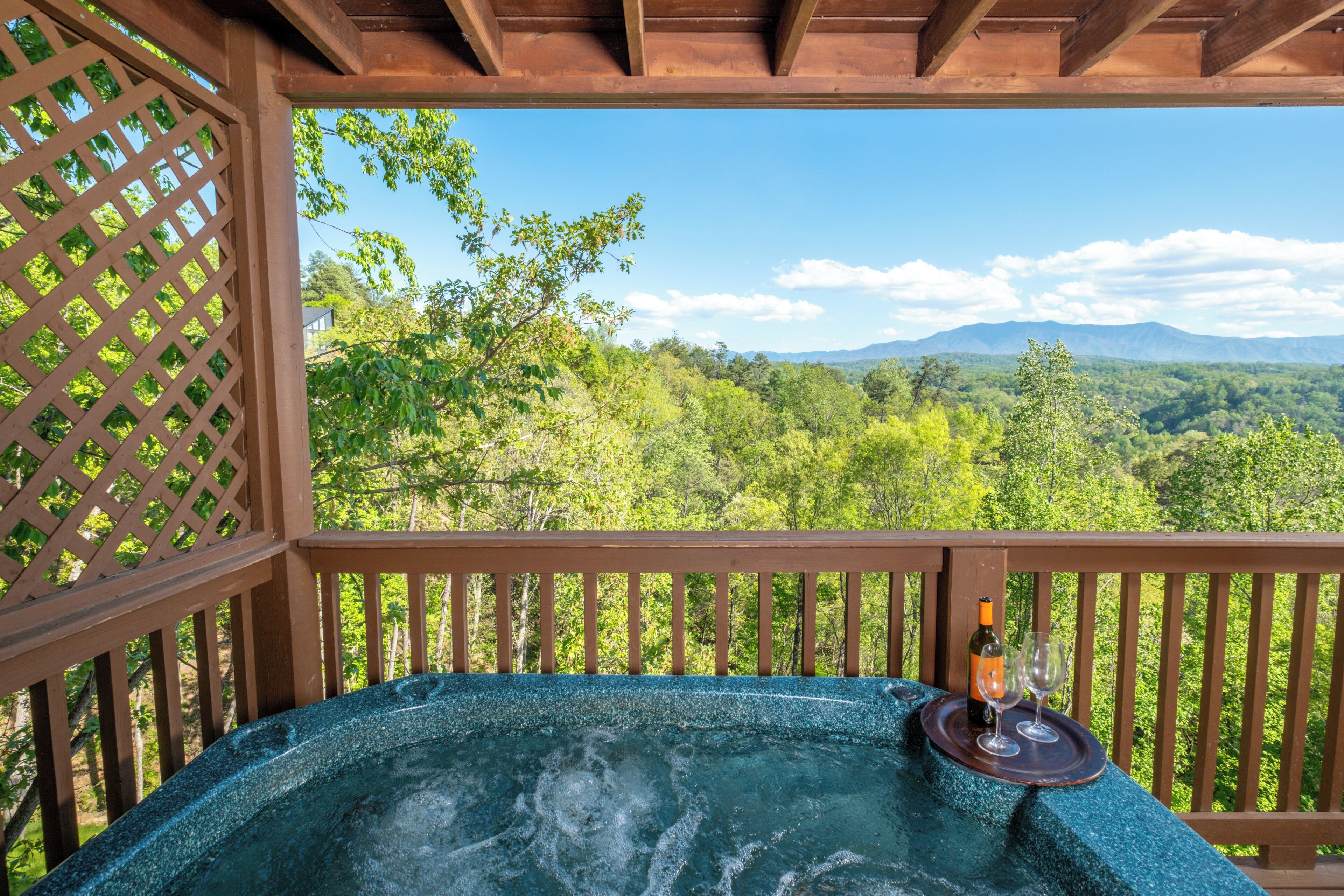 Un jacuzzi con una botella y copas sobre una terraza de madera, con vistas a un bosque y montañas.