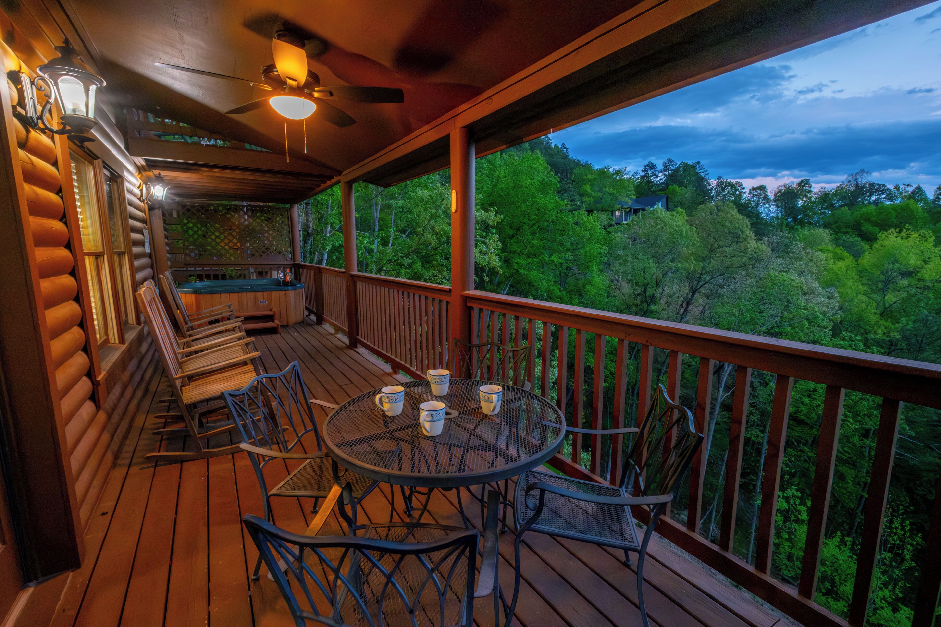 A wooden deck with a round table and chairs, a ceiling fan, and a view of a forested landscape.