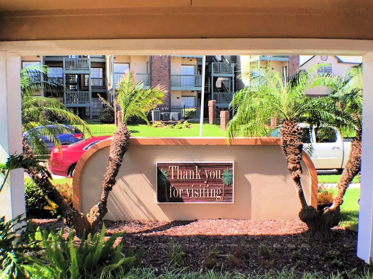 A sign with "Thank you for visiting" in a landscaped area with palm trees and a building in the background.