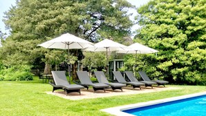 Un área junto a la piscina con sillones de descanso y sombrillas, rodeada de abundante vegetación.
