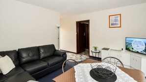 A living room with a black leather sofa, a dining table with a white lace table runner, a television on the wall, and a framed picture above the TV. - SELF CONTAINED-1 Bdrm Apartments (Albany)