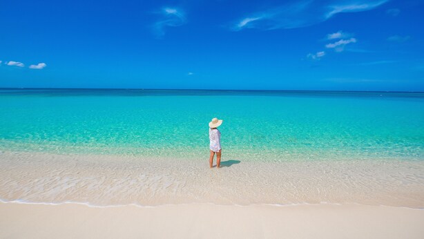 En person som står på en strand med kristallklart turkost vatten och en klar himmel.