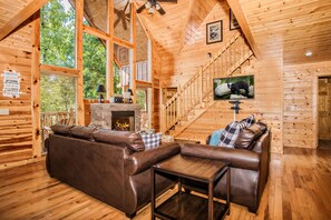 A wooden cabin interior with a leather sofa, a fireplace, a staircase, and a large window overlooking trees.