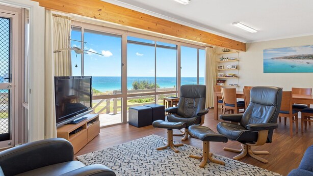 A spacious living room with a large TV, comfortable seating, a wooden floor, and a view of the ocean through large windows. - Sand Castles - 34 Gold Coast Drive (CARRICKALINGA)