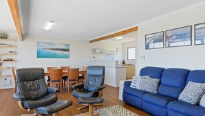 A modern living room with a blue sofa, leather chairs, wooden dining table, and a kitchen in the background. - Sand Castles - 34 Gold Coast Drive (CARRICKALINGA)