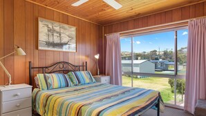 A bedroom with a bed, a nightstand, a lamp, a window with curtains, and a painting on the wall. - Sand Castles - 34 Gold Coast Drive (CARRICKALINGA)