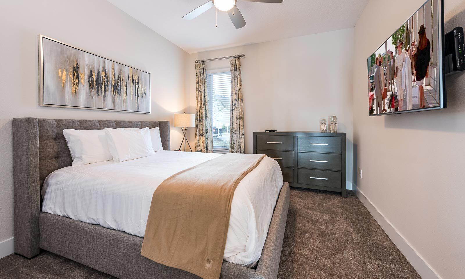 A bedroom with a bed, a dresser, a ceiling fan, a window with curtains, and a painting on the wall.
