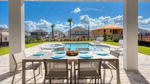 Un área de comedor al aire libre con piscina, mesas dispuestas con platos y vasos azules, y vistas a edificios residenciales al fondo.