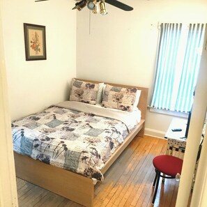 A bedroom with a bed, a ceiling fan, a window with curtains, and a small table with a chair.