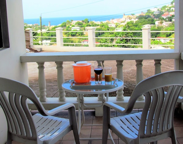 A balcony with two chairs, a table with drinks, and a view of a coastal town.