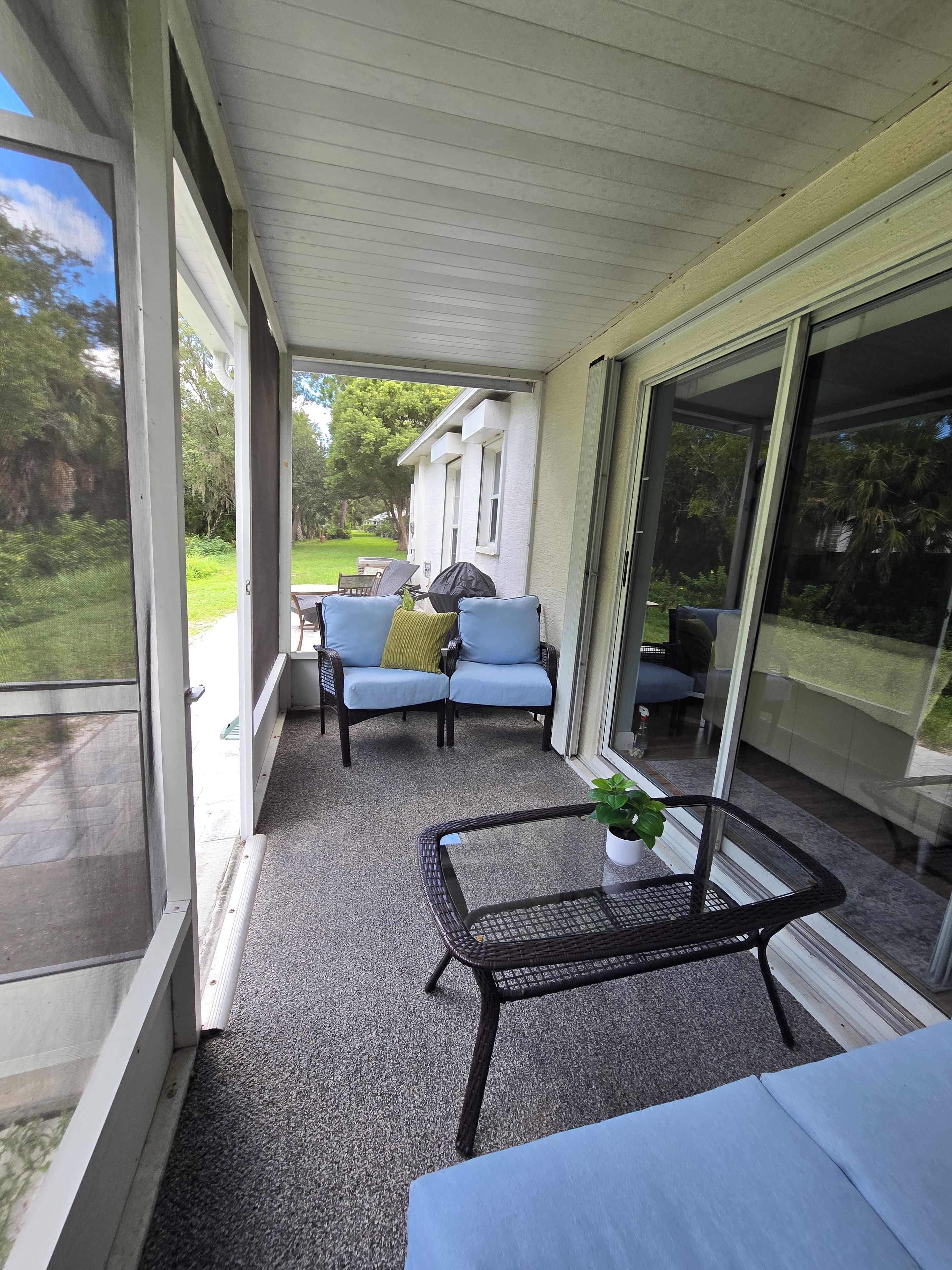 A covered outdoor seating area with a table and chairs, a potted plant, and a view of a house and greenery.