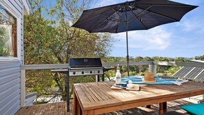 Une terrasse extérieure avec un barbecue, une table sur laquelle se trouvent une bouteille et un chapeau, et une vue sur les arbres et les collines.