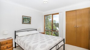 Une chambre à coucher comprenant un lit, une table de chevet, une commode, une armoire en bois, une fenêtre donnant sur des arbres et un tableau encadré accroché au mur.