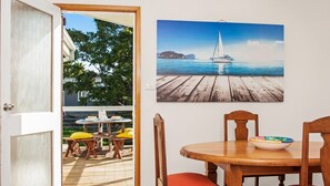 An open door leading to a patio with a wooden table and chairs, a painting of a sailboat on water, and a view of a house and trees. - Mollymook Sun Sand Surf (Mollymook)