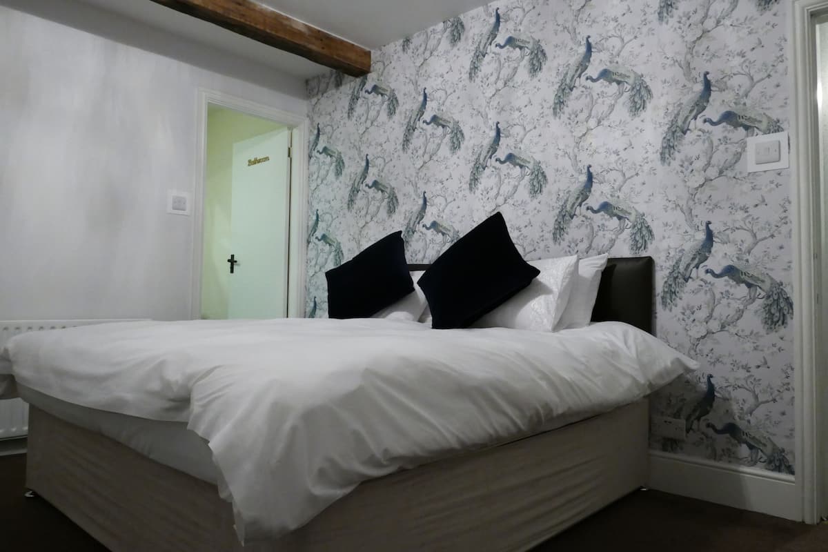 A bedroom with a bed, white bedding, black pillows, and a wall with a peacock pattern.