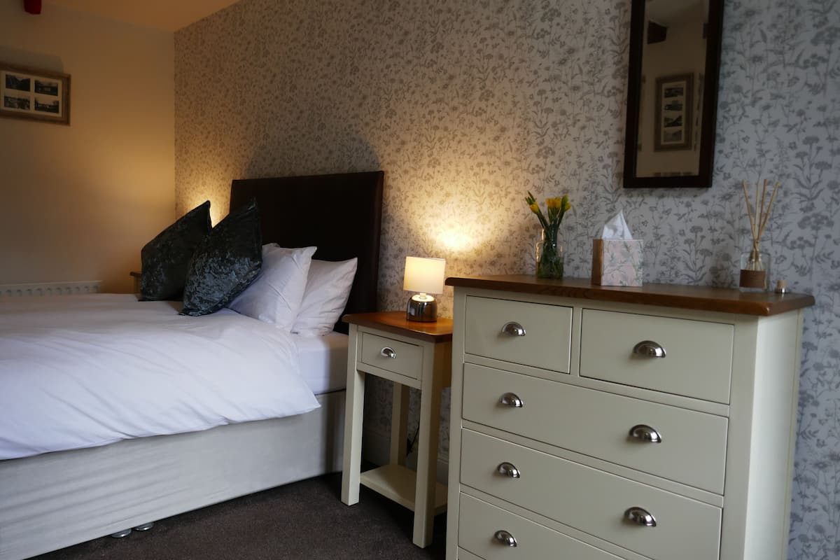 A neatly made bed with white linens and a dark headboard, a bedside table with a lamp, and a dresser with drawers.
