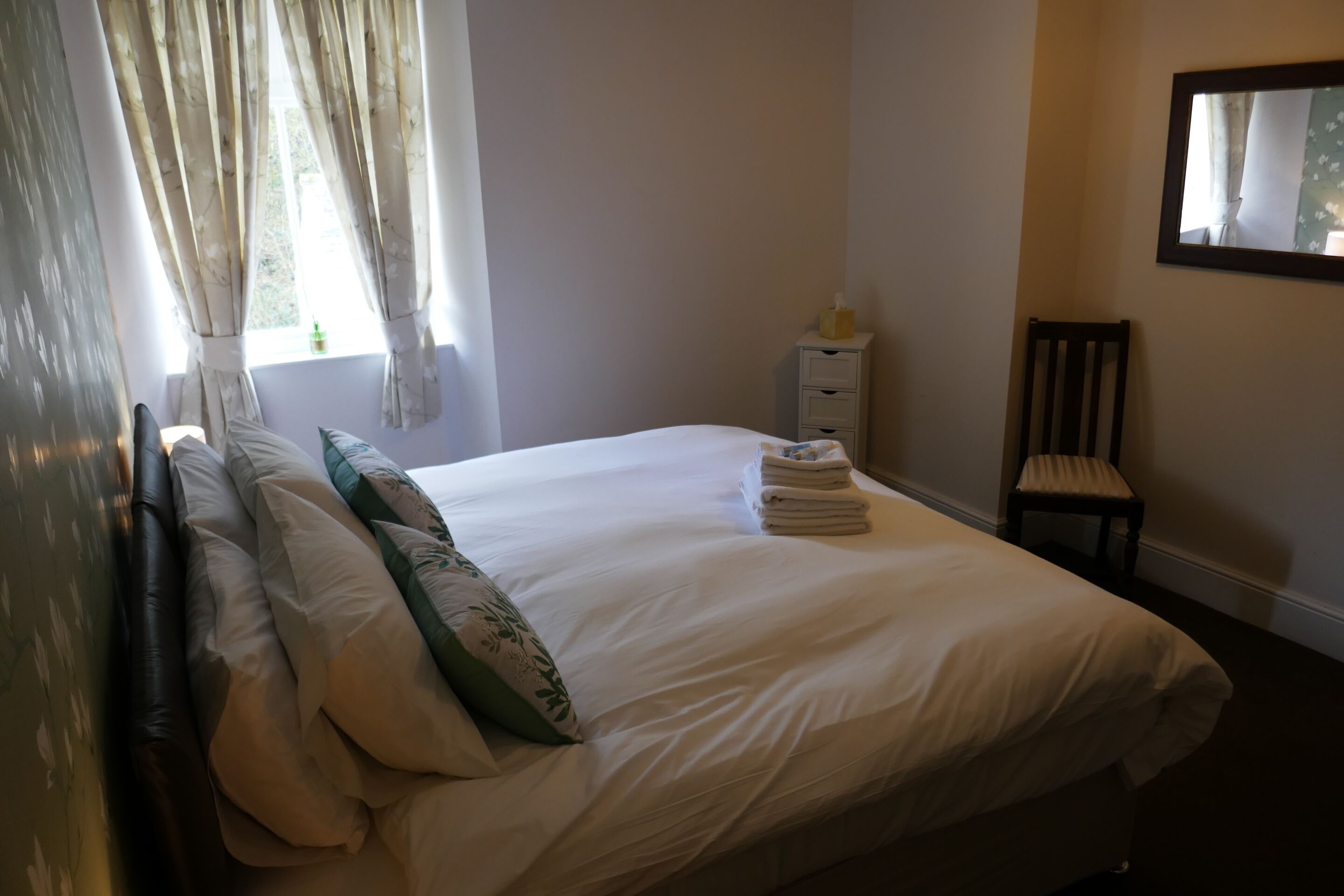Un lit bien fait, avec du linge de lit blanc et un coussin vert, une petite table de chevet avec une boîte de mouchoirs, une chaise et une fenêtre avec des rideaux.