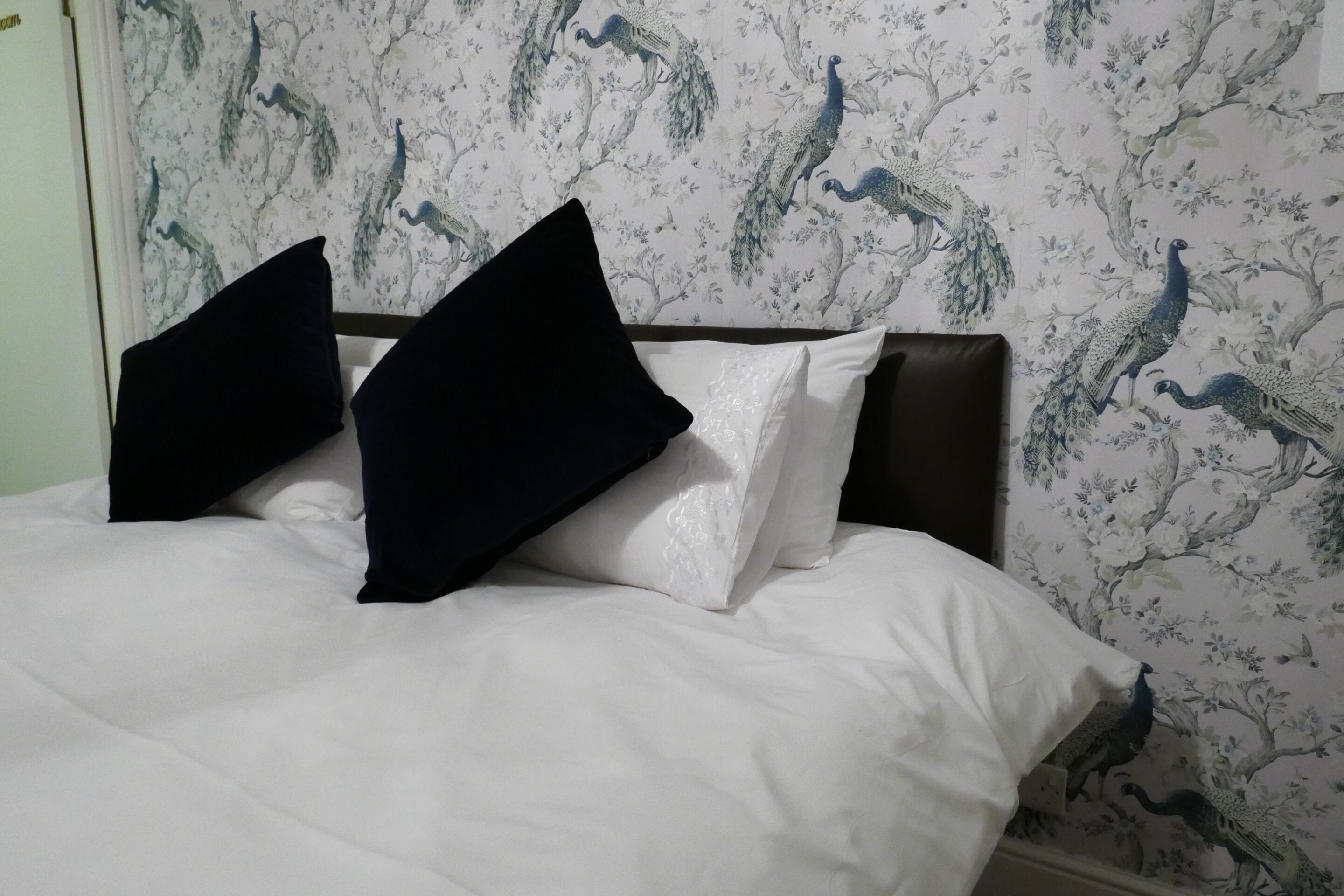 Una cama con ropa de cama blanca y almohadas blancas y negras, frente a una pared con papel tapiz de pavos reales y motivos florales.