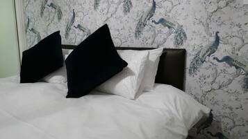 Una cama con ropa de cama blanca y almohadas blancas y negras, frente a una pared con papel tapiz de pavos reales y motivos florales.