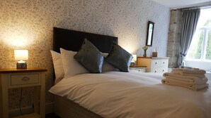 A neatly made bed with pillows, a bedside lamp, a stack of towels, and a nightstand with a drawer. - The Victoria (Skipton)