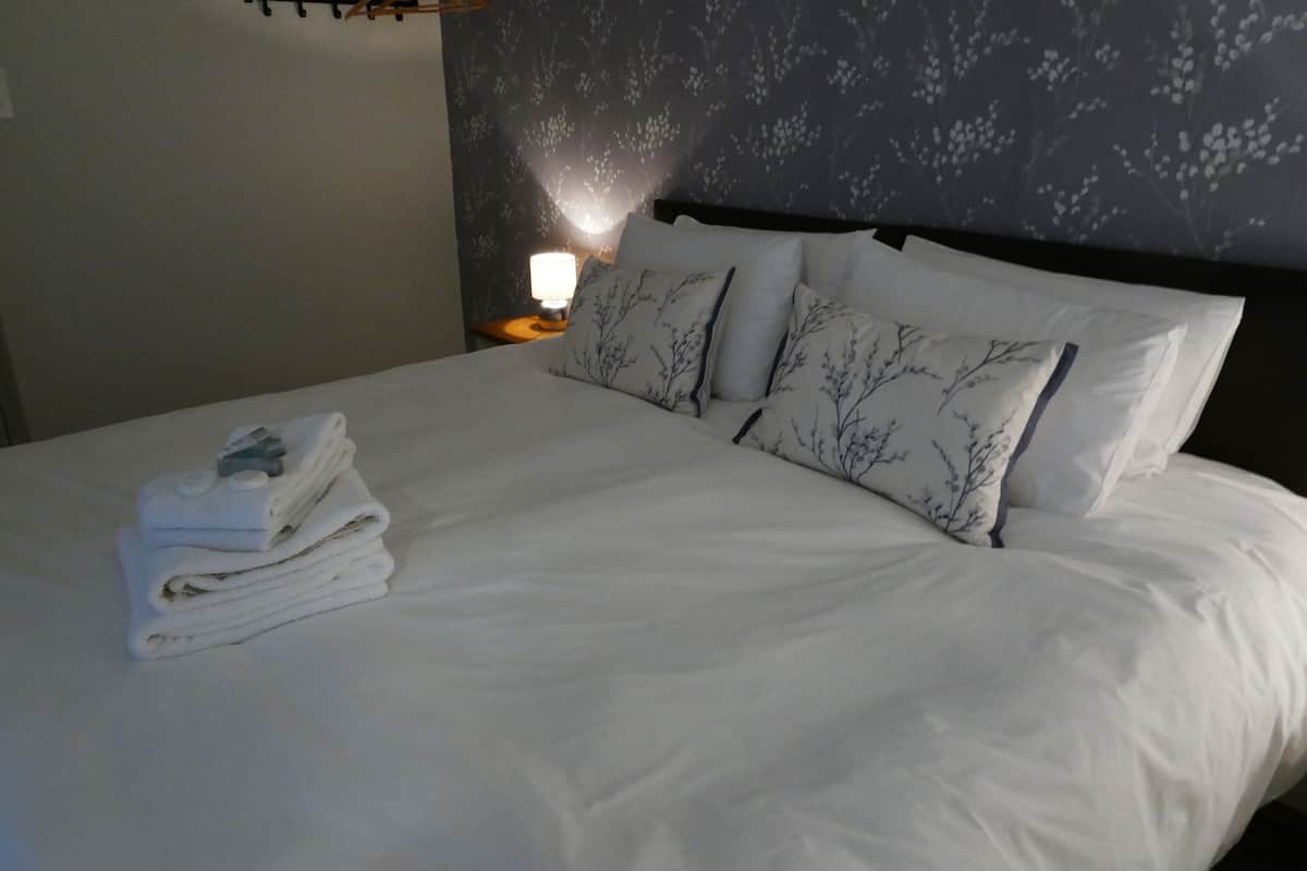 A neatly made bed with white linens and tree-patterned pillows, a bedside lamp, and a stack of folded towels.