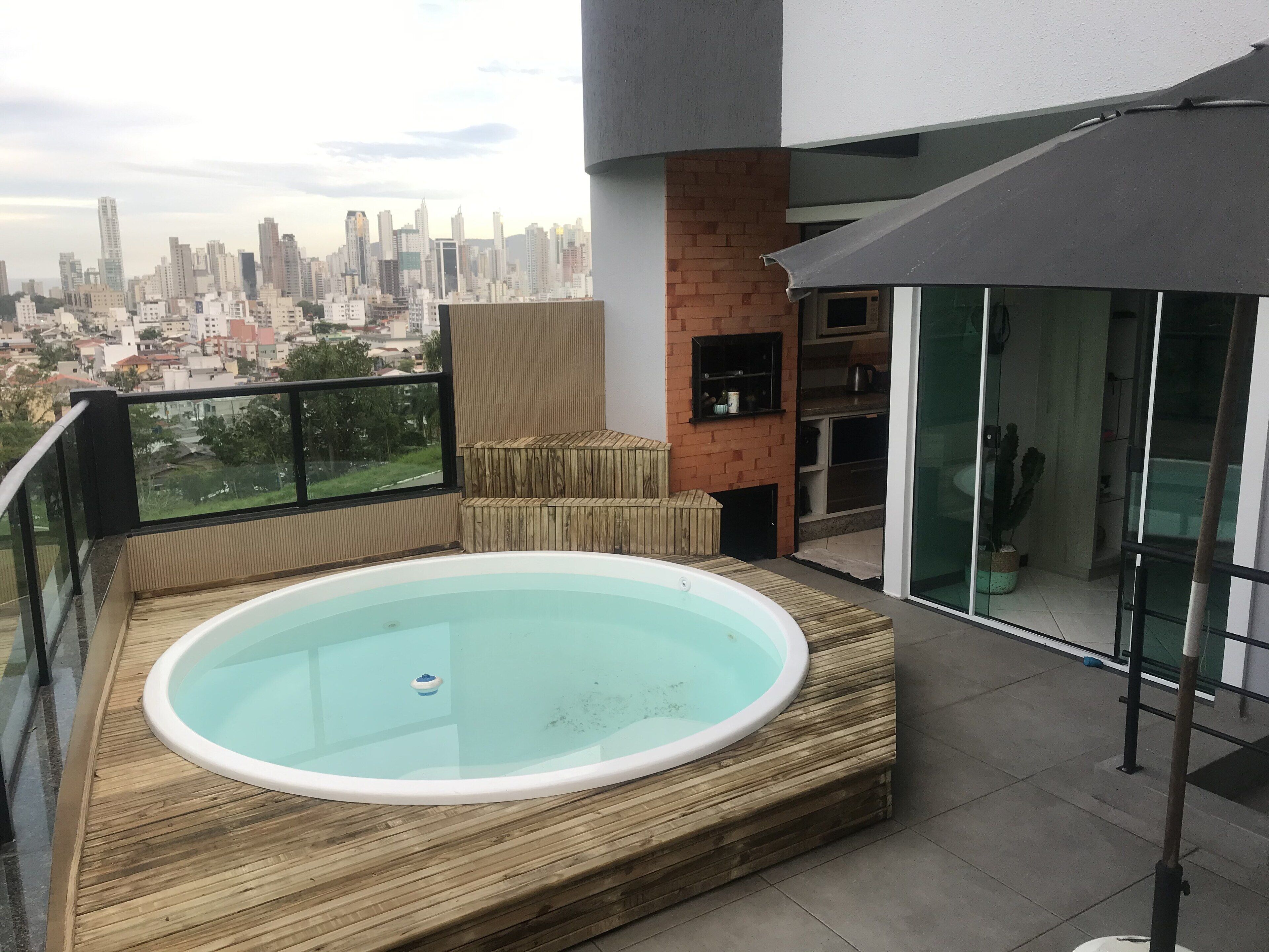 A rooftop terrace with a hot tub, a glass balustrade, a brick wall, and a city view.