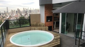 Eine Dachterrasse mit Whirlpool, Glasgeländer, Backsteinmauer und Blick auf die Stadt.