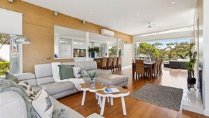 A spacious living room with a sofa, coffee table, and dining area with chairs. There is a kitchen in the background with cabinets and a dining table. - Fairway View (FLINDERS)