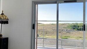 Un dormitorio con cama, mesita de noche y lámpara. Hay un amplio ventanal corredero que da a un balcón exterior con vistas a un paisaje montañoso.